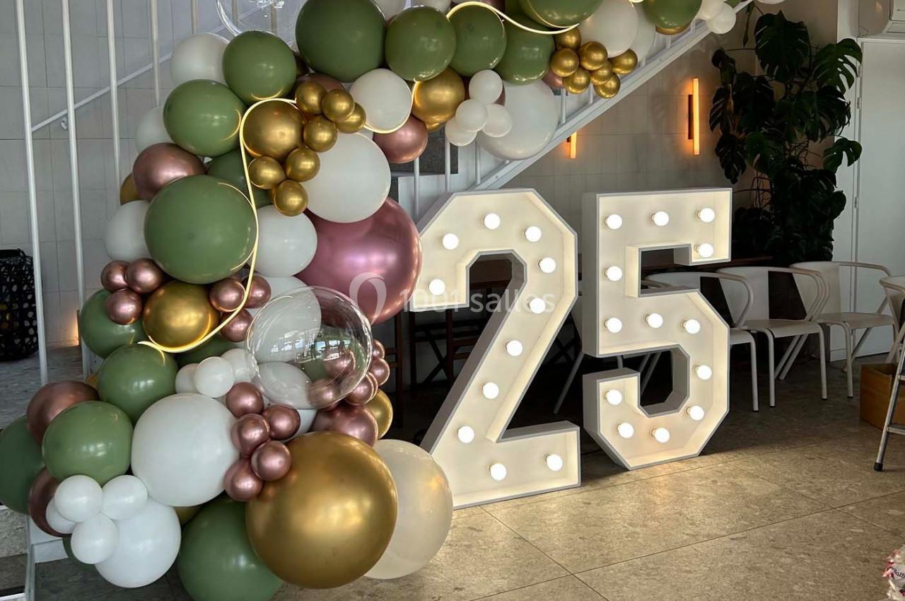Décoration festive avec ballons verts, blancs et dorés entourant un chiffre lumineux ’25’ sous un escalier.