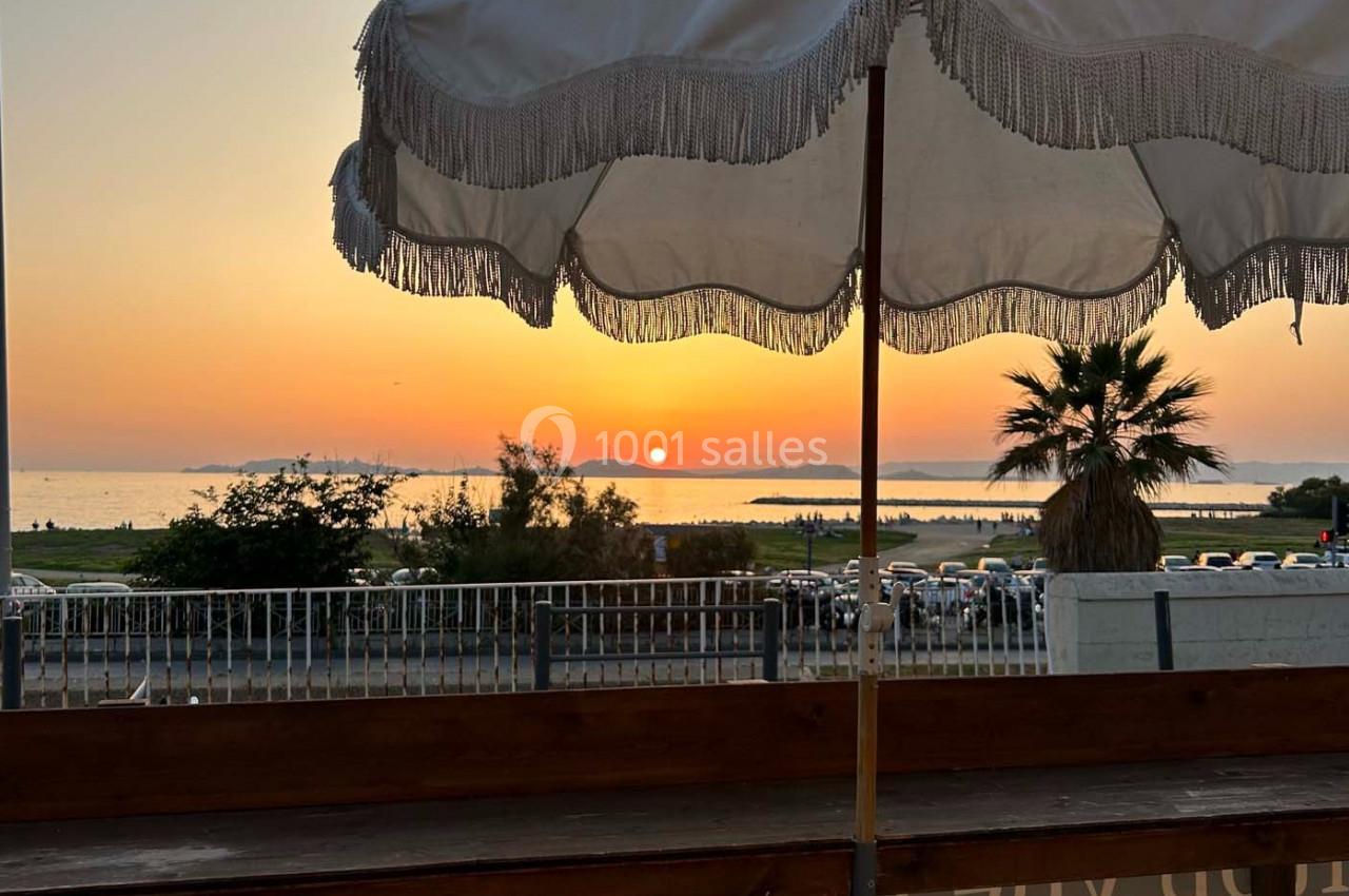 Vue d'un coucher de soleil sur la mer, avec un parasol blanc au premier plan et des palmiers en arrière-plan.