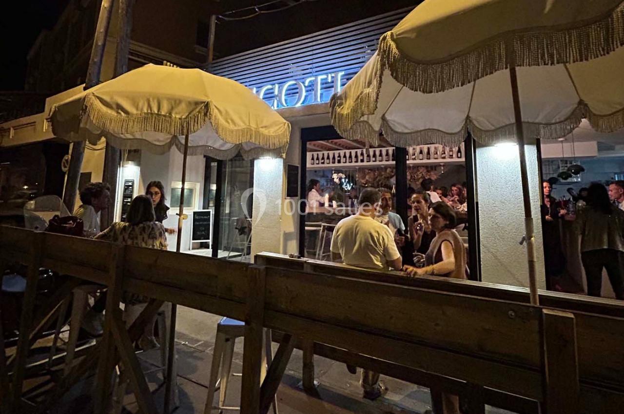 Terrasse d'un bar animé en soirée, avec des parasols éclairés et des clients discutant autour de tables en bois.