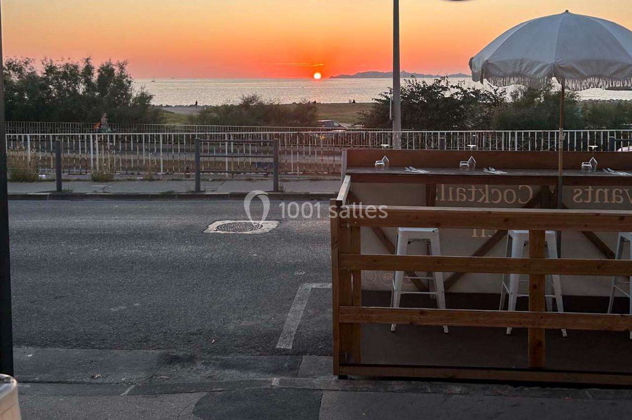 Vue d'une terrasse en bord de route avec des tabourets, face à un coucher de soleil sur la mer.
