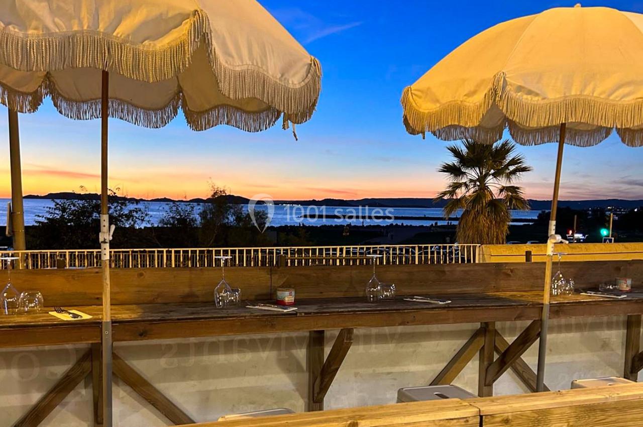 Terrasse en bois avec parasols, vue sur un coucher de soleil au bord de l'eau et palmiers à l'horizon.