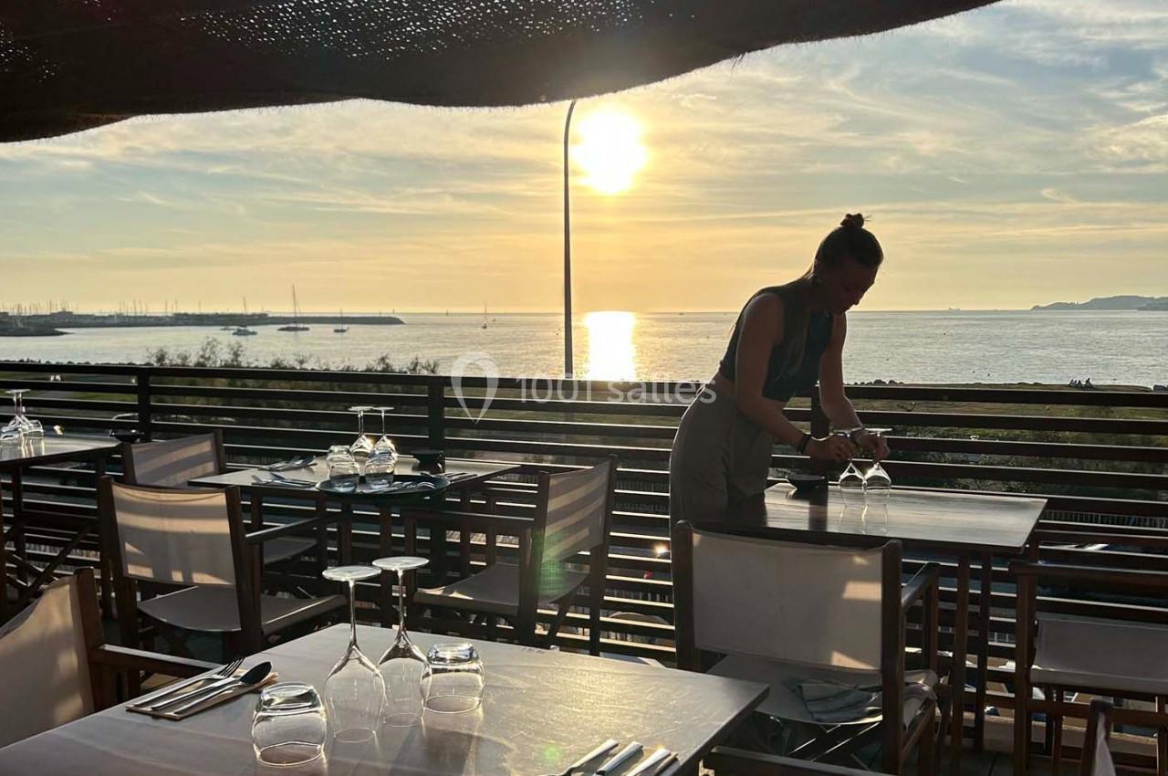 Une serveuse prépare une table en terrasse avec vue sur la mer au coucher du soleil.