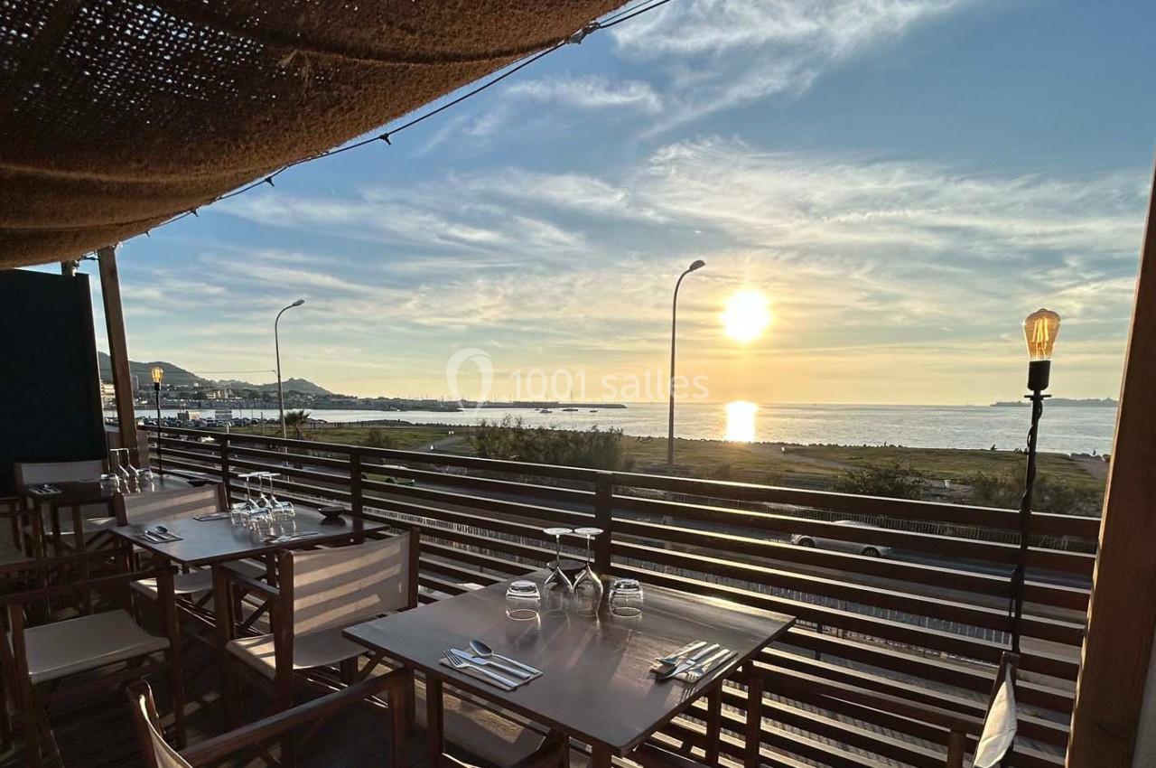 Terrasse en bois d'un restaurant avec vue sur la mer au coucher du soleil, tables dressées et ciel dégagé.