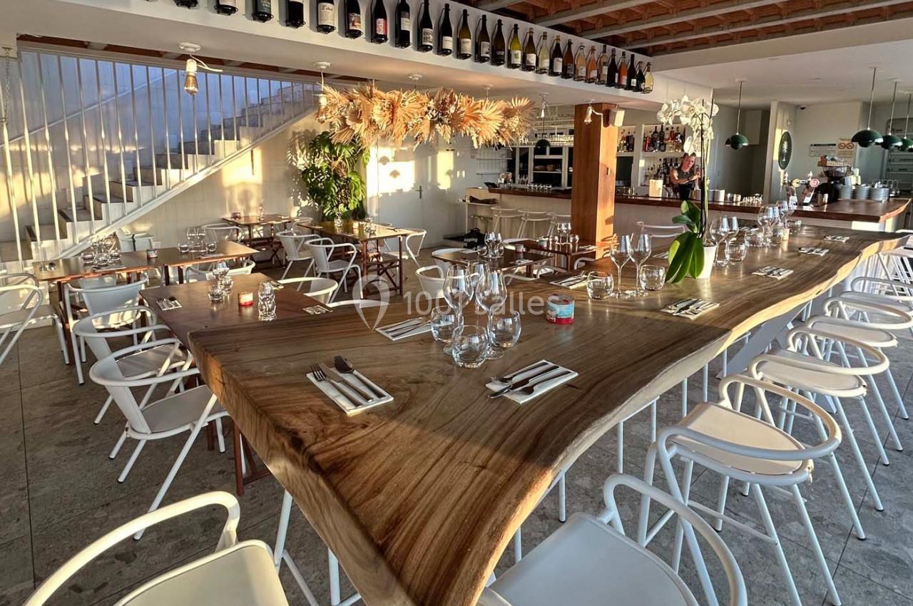Salle de restaurant lumineuse avec une grande table en bois naturel, chaises blanches et décoration suspendue.
