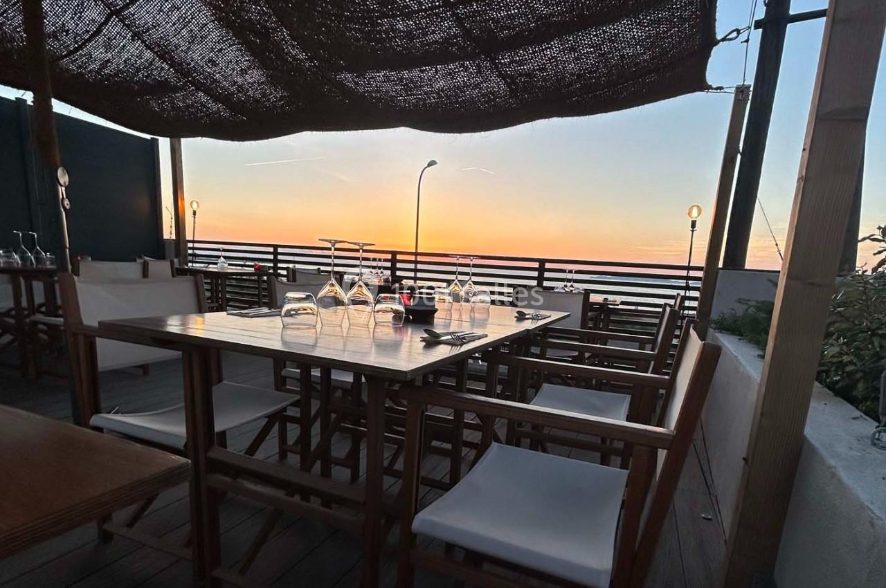 Table dressée en terrasse avec vue sur un coucher de soleil, chaises en bois et lumière tamisée.