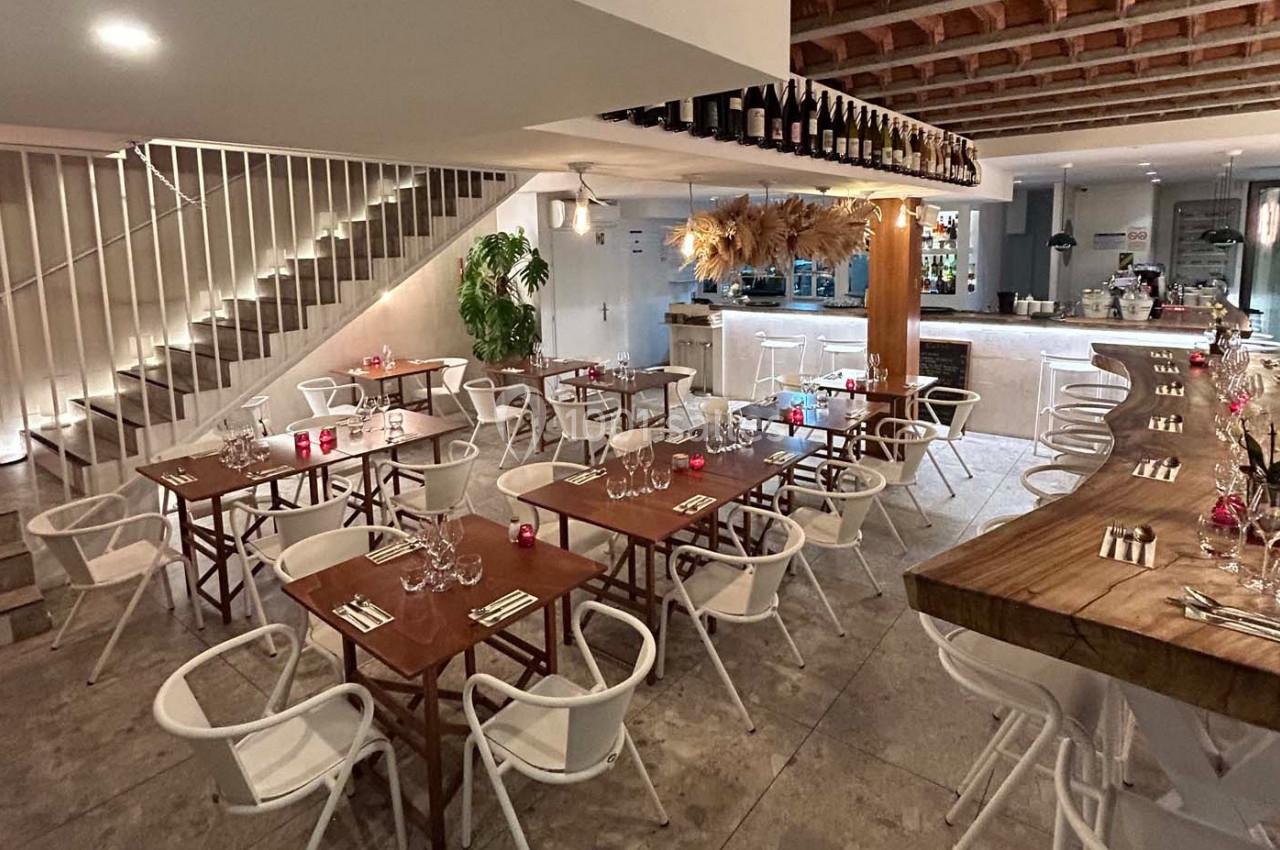 Salle de restaurant moderne avec tables en bois, chaises blanches, bar en arrière-plan et décoration suspendue.