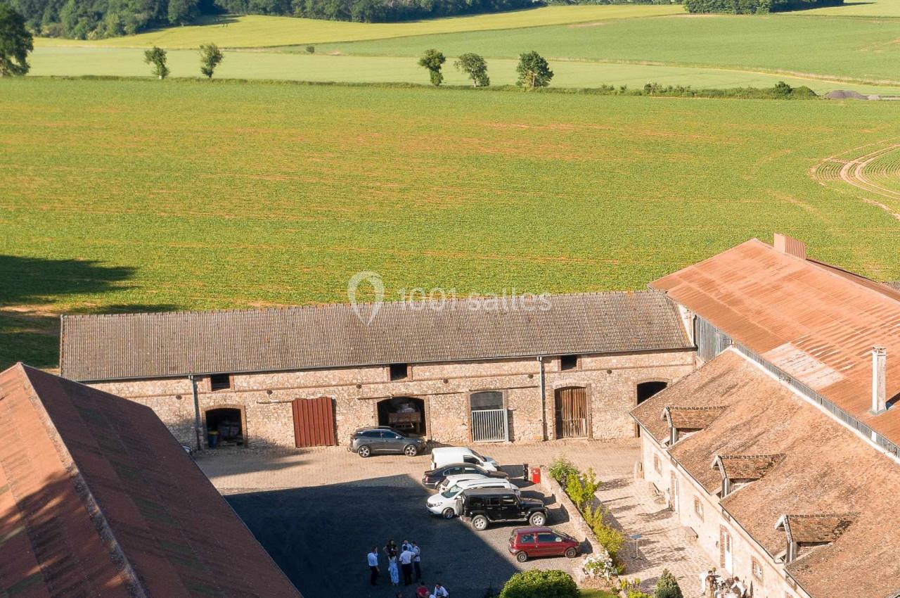 Vue aérienne d'une ferme en pierre entourée de champs verdoyants, avec des personnes rassemblées dans la cour.