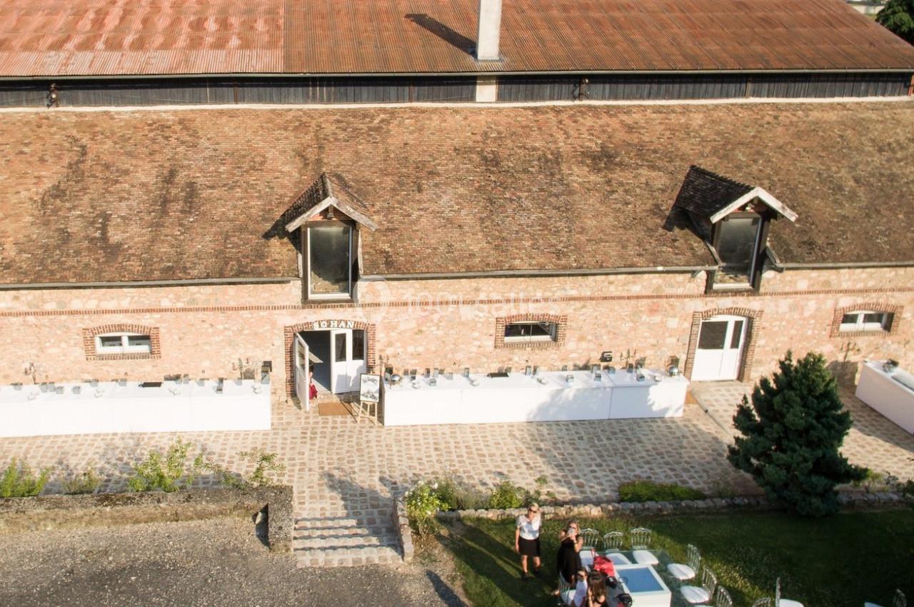 Bâtiment en briques avec toit en tuiles, petites fenêtres sous les combles, et terrasse aménagée avec des personnes.