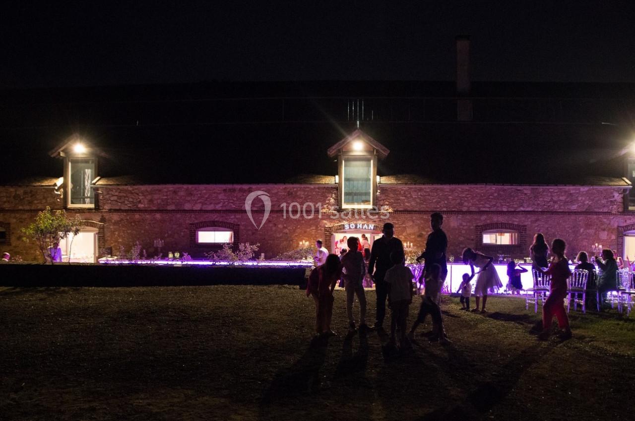 Des personnes discutent devant un bâtiment en pierre éclairé, de nuit, avec des lumières violettes au sol.