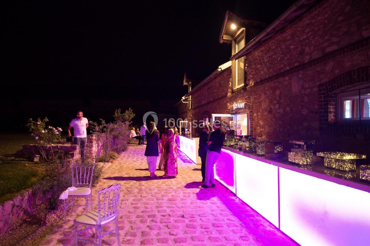 Personnes discutant près d'un bar éclairé en violet, situé à l'extérieur d'un bâtiment en pierre, de nuit.