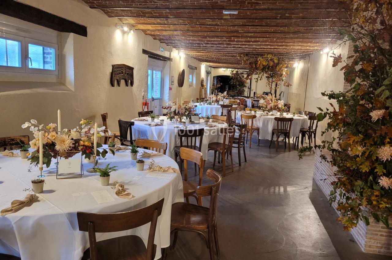 Salle décorée pour un événement avec des tables rondes dressées, ornées de fleurs et de bougies, sous un plafond en bois.