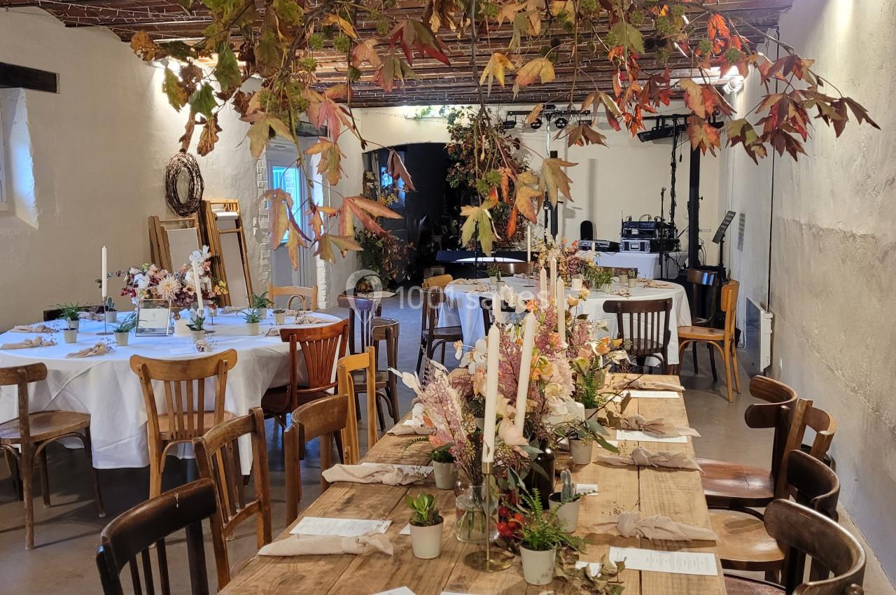 Salle décorée pour un événement, avec une grande table en bois ornée de bougies et de fleurs, sous un plafond rustique.