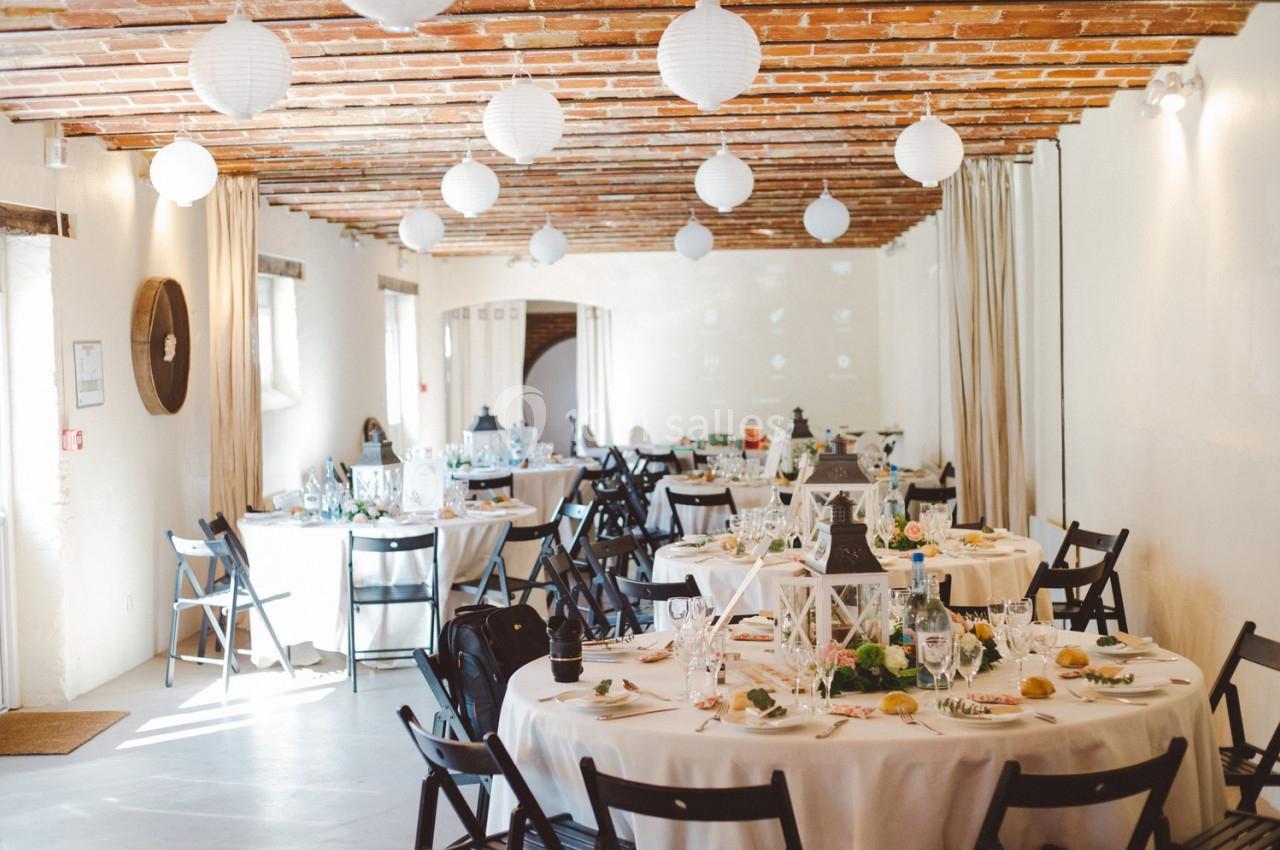 Salle de réception décorée avec des tables rondes, nappes blanches, chaises noires et lanternes suspendues au plafond.