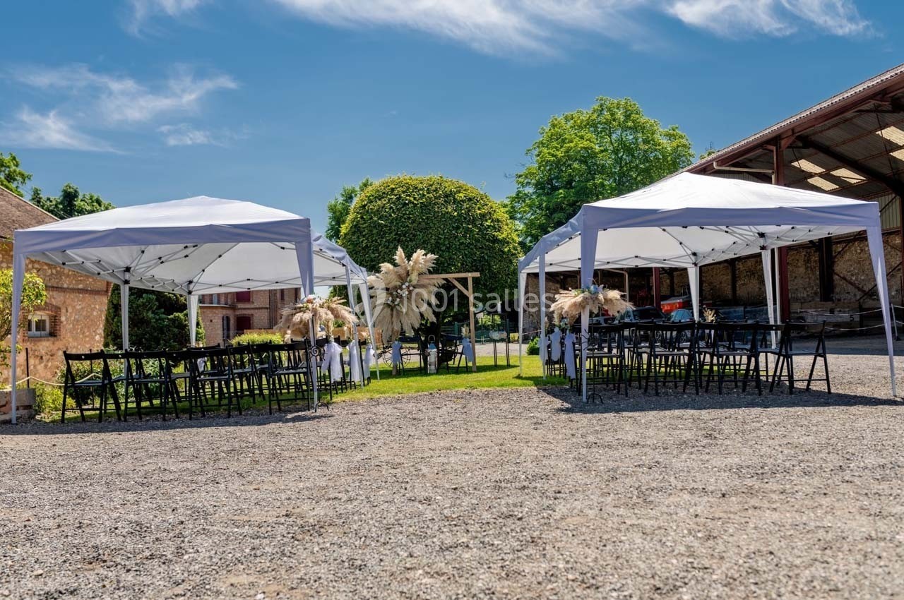 Deux tentes blanches installées en extérieur avec des chaises noires, entourées de verdure et de bâtiments rustiques.