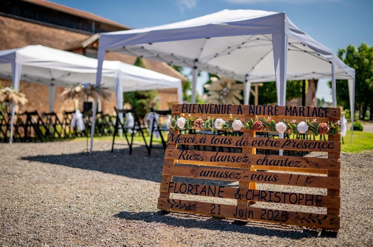 Panneau en bois décoré de fleurs annonçant un mariage, placé devant des tentes blanches en extérieur.