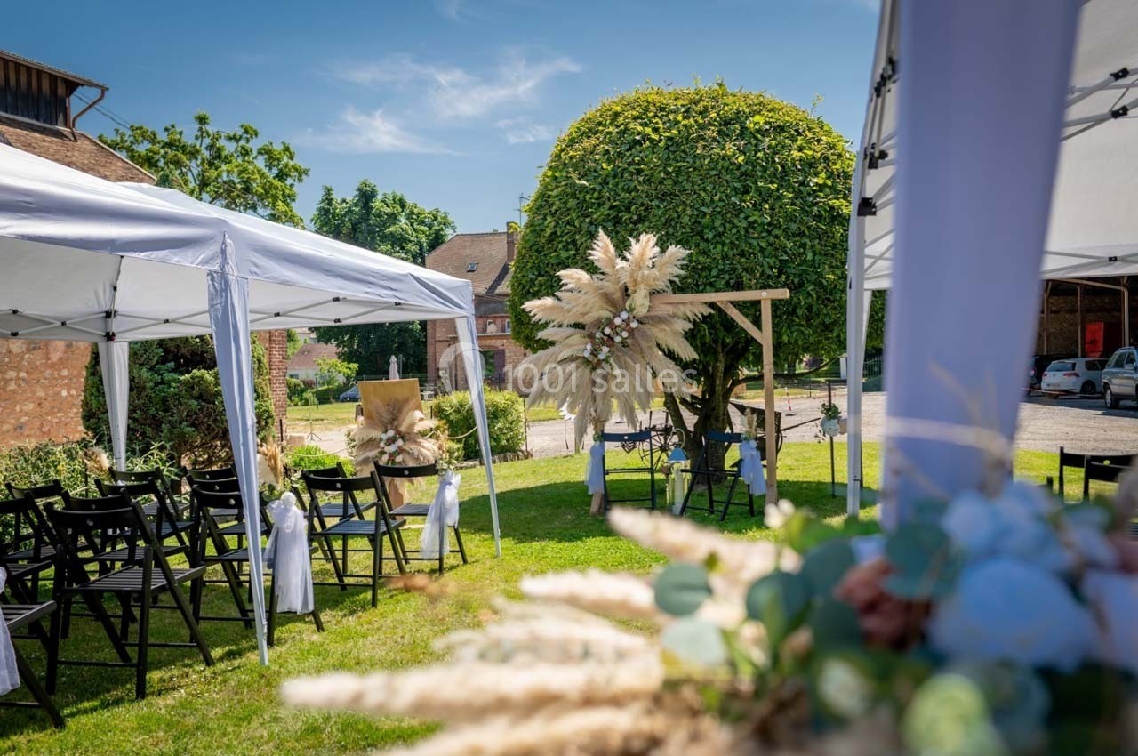Cérémonie en plein air avec chaises noires, tonnelles blanches et décorations florales sous un arbre taillé.