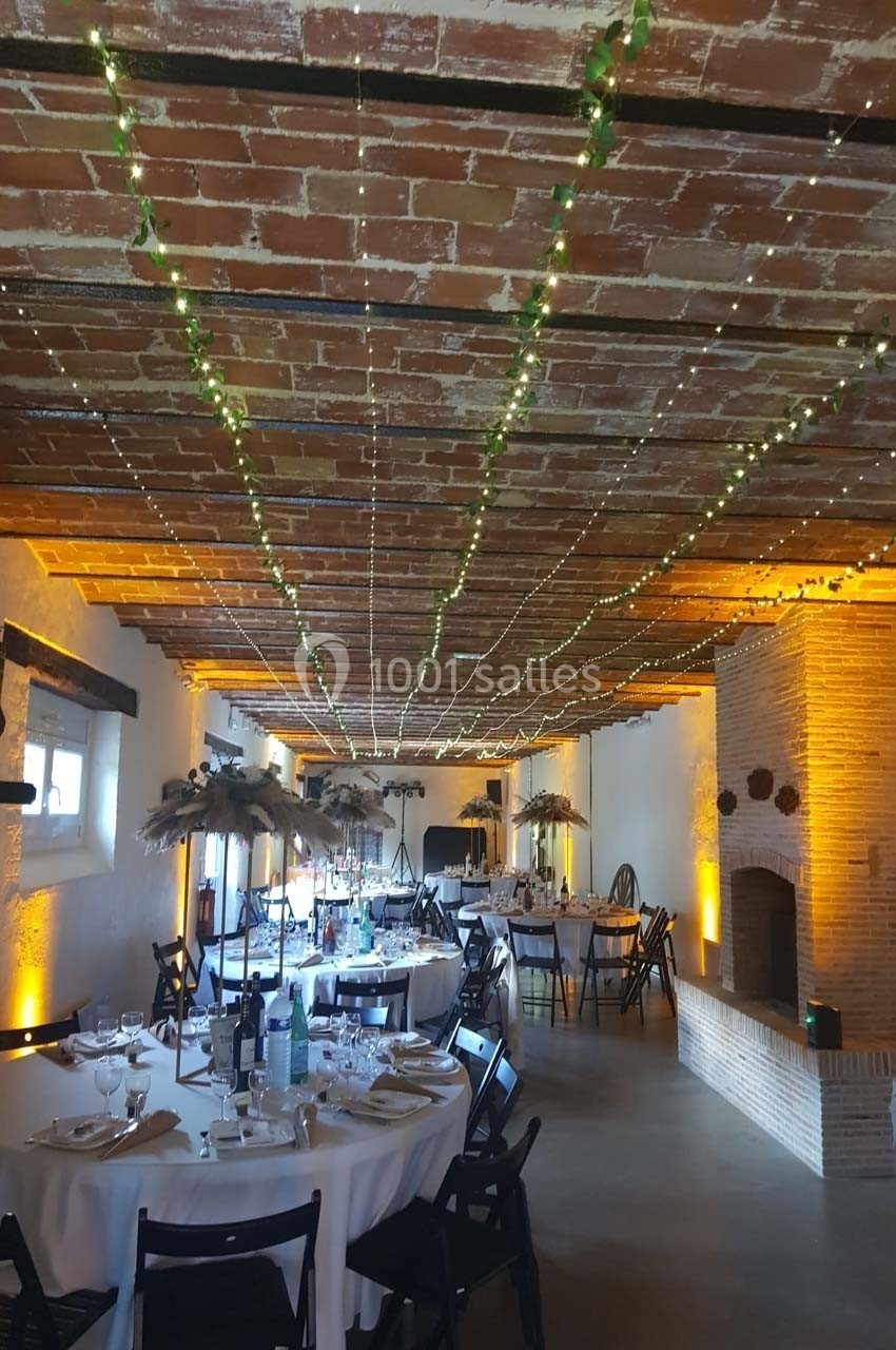 Salle de réception avec tables rondes dressées, plafond en briques décoré de guirlandes lumineuses et cheminée en briques.