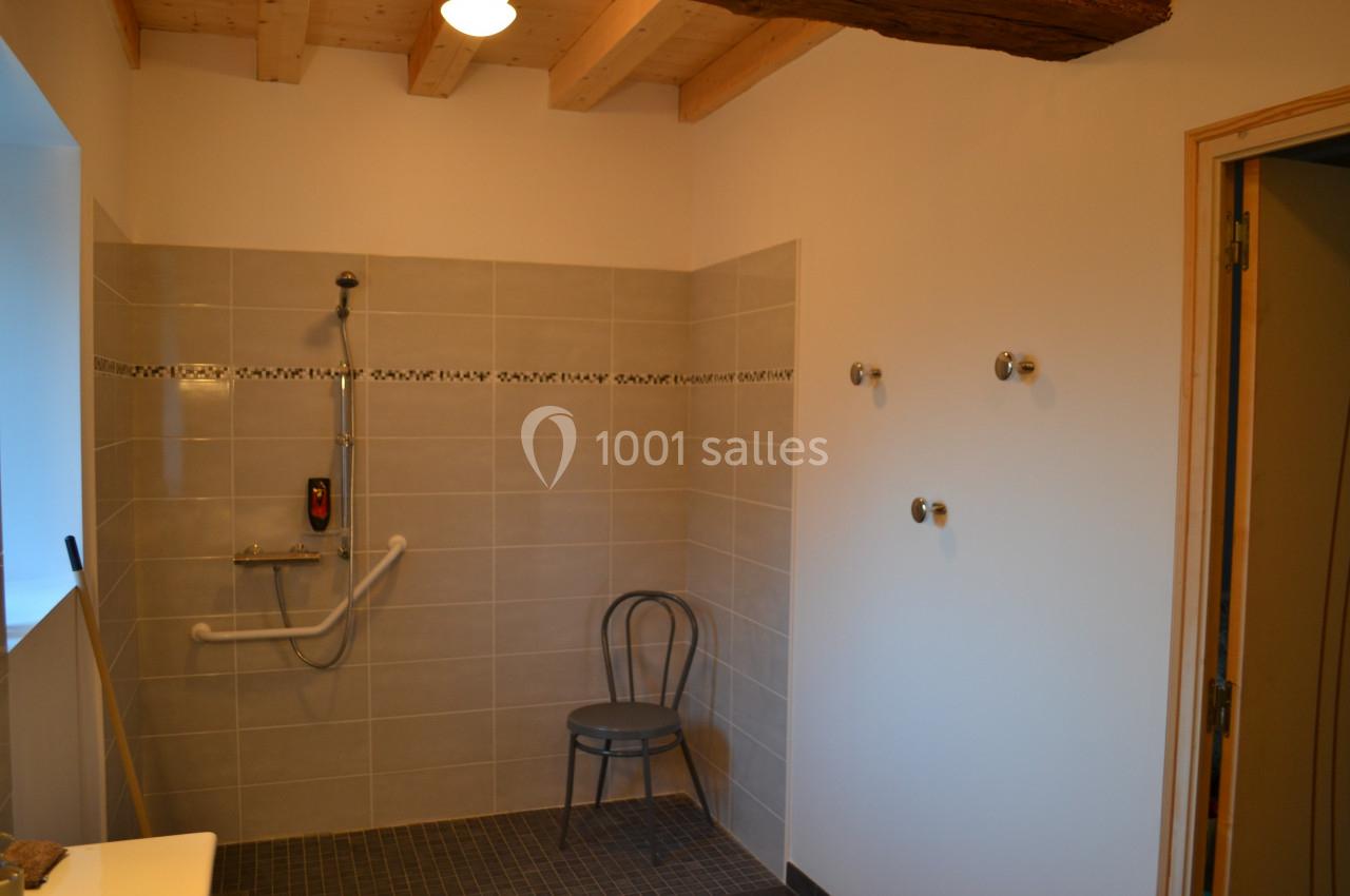 Salle de bain avec douche à l'italienne, barre d'appui, chaise et carrelage beige, éclairée par une lumière naturelle.