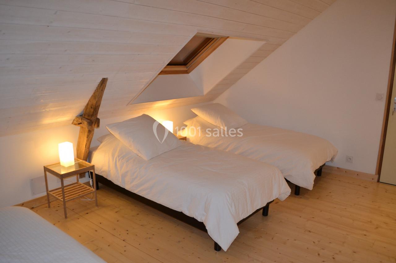 Trois lits simples avec draps blancs dans une chambre mansardée au plafond en bois clair, éclairée par des lampes de chevet.