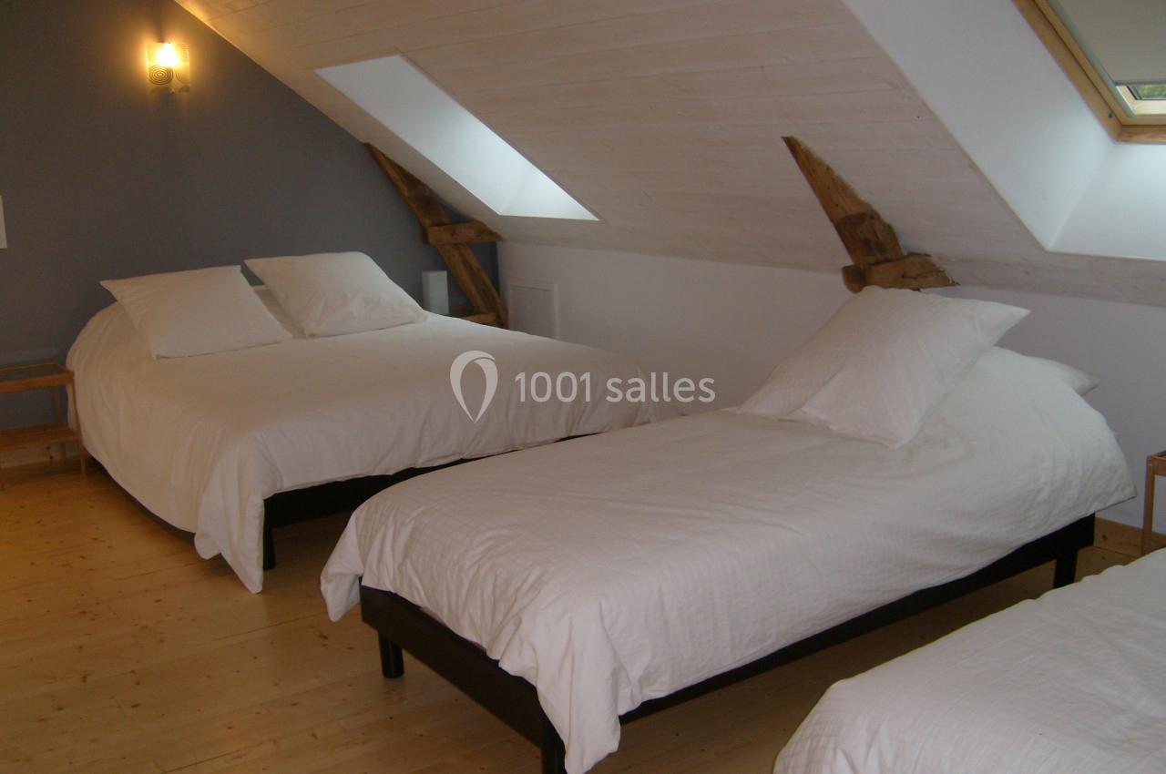 Trois lits avec draps blancs dans une chambre mansardée au parquet en bois clair, éclairée par une fenêtre de toit.