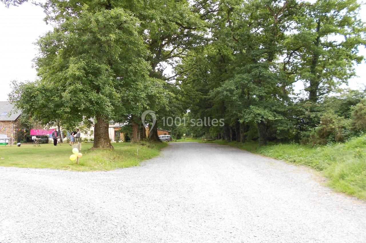 Chemin de gravier bordé d'arbres, avec des bâtiments et des personnes visibles sur la gauche.