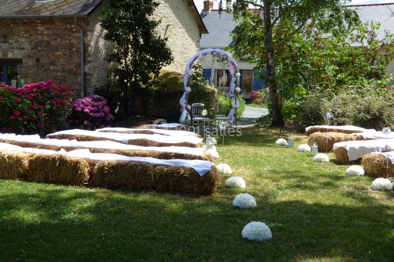 Chaises et bottes de paille décorées disposées en extérieur pour une cérémonie, avec une arche fleurie en arrière-plan.