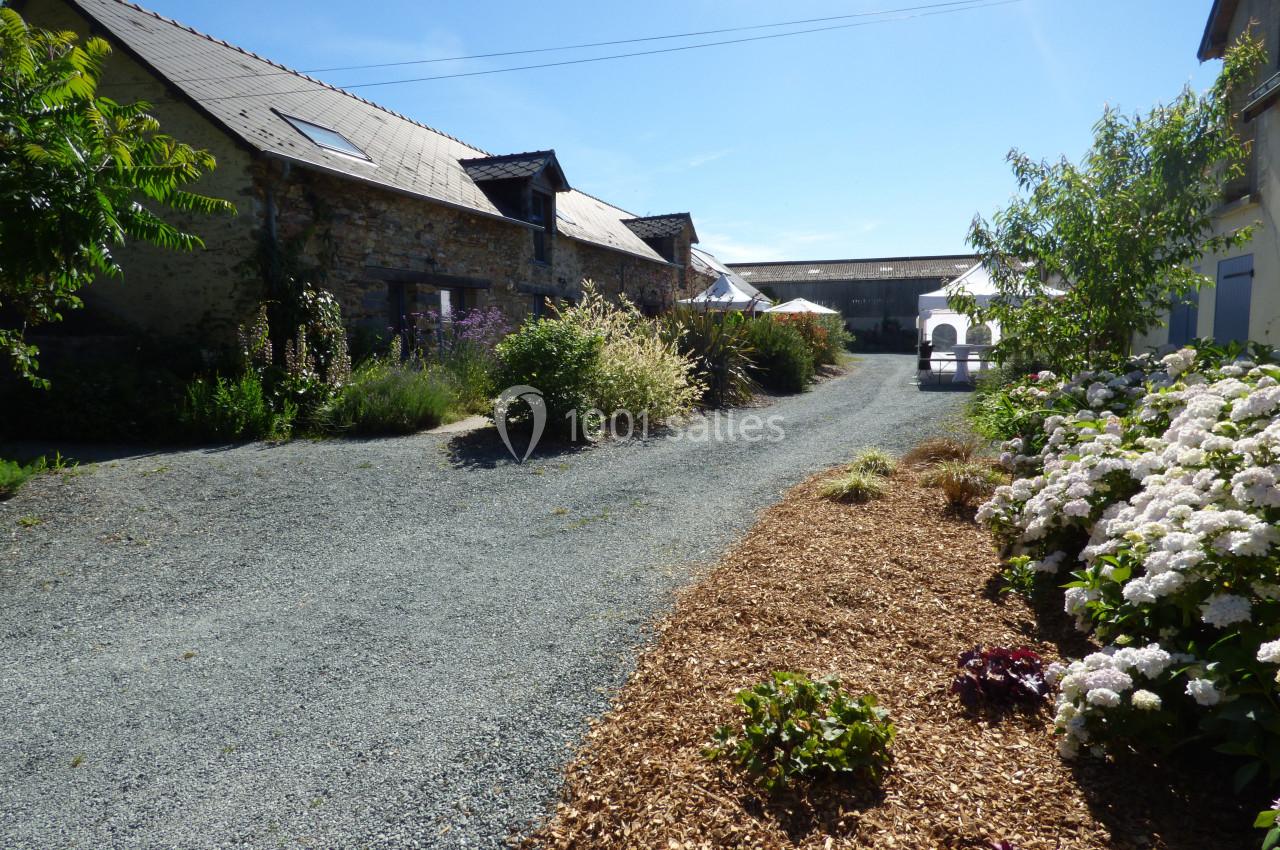 Cour en gravier bordée de fleurs, arbustes et bâtiments en pierre sous un ciel bleu clair.