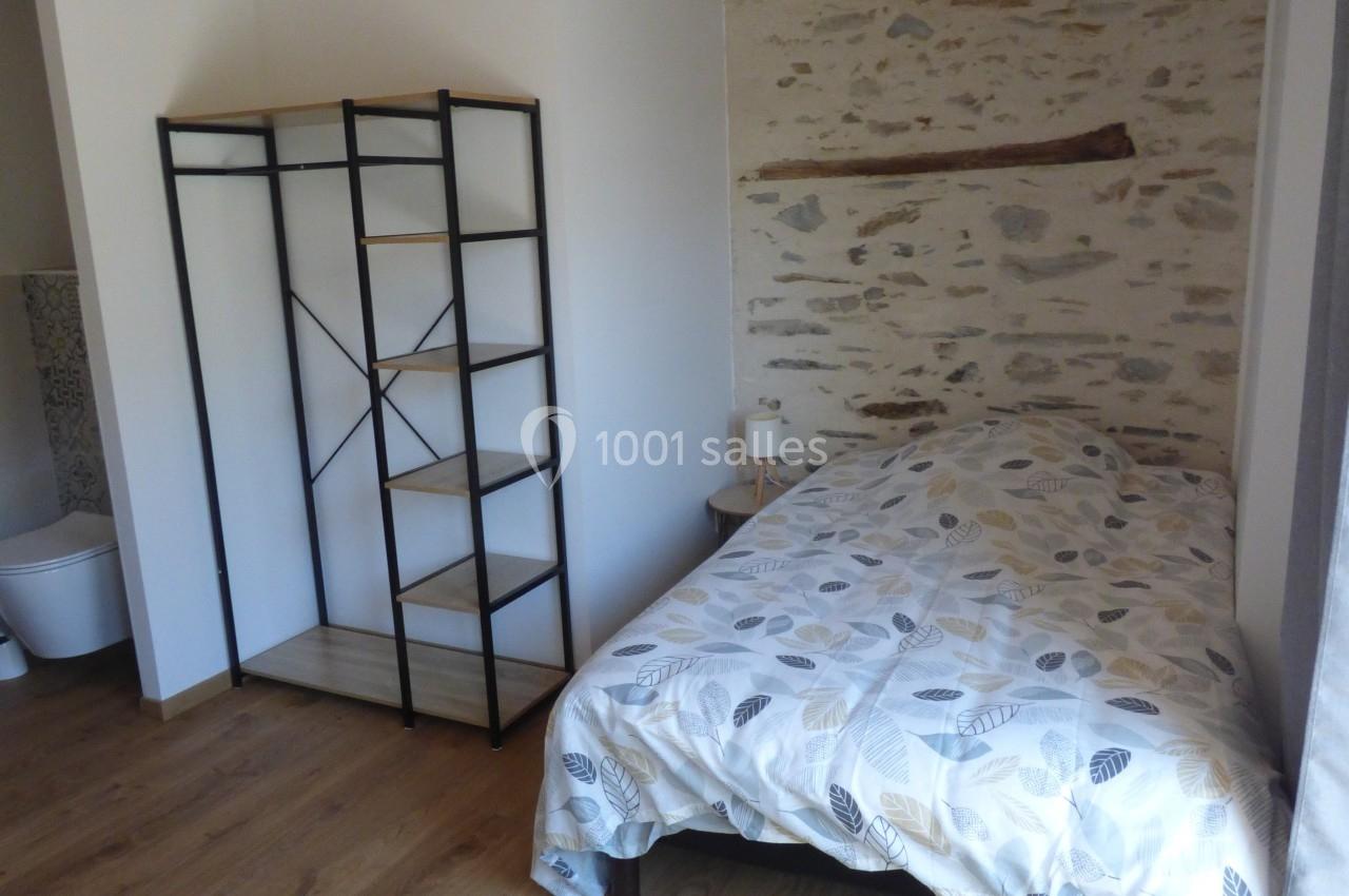 Chambre avec lit simple, étagère en métal noir, mur en pierre apparente et plafond en bois.