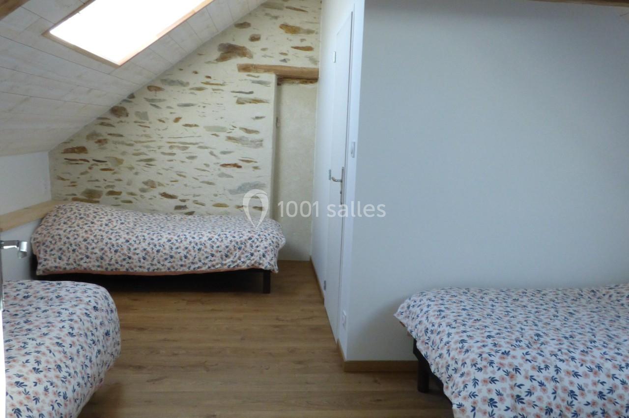 Chambre mansardée avec trois lits simples, parquet au sol, murs clairs et poutres apparentes en bois.