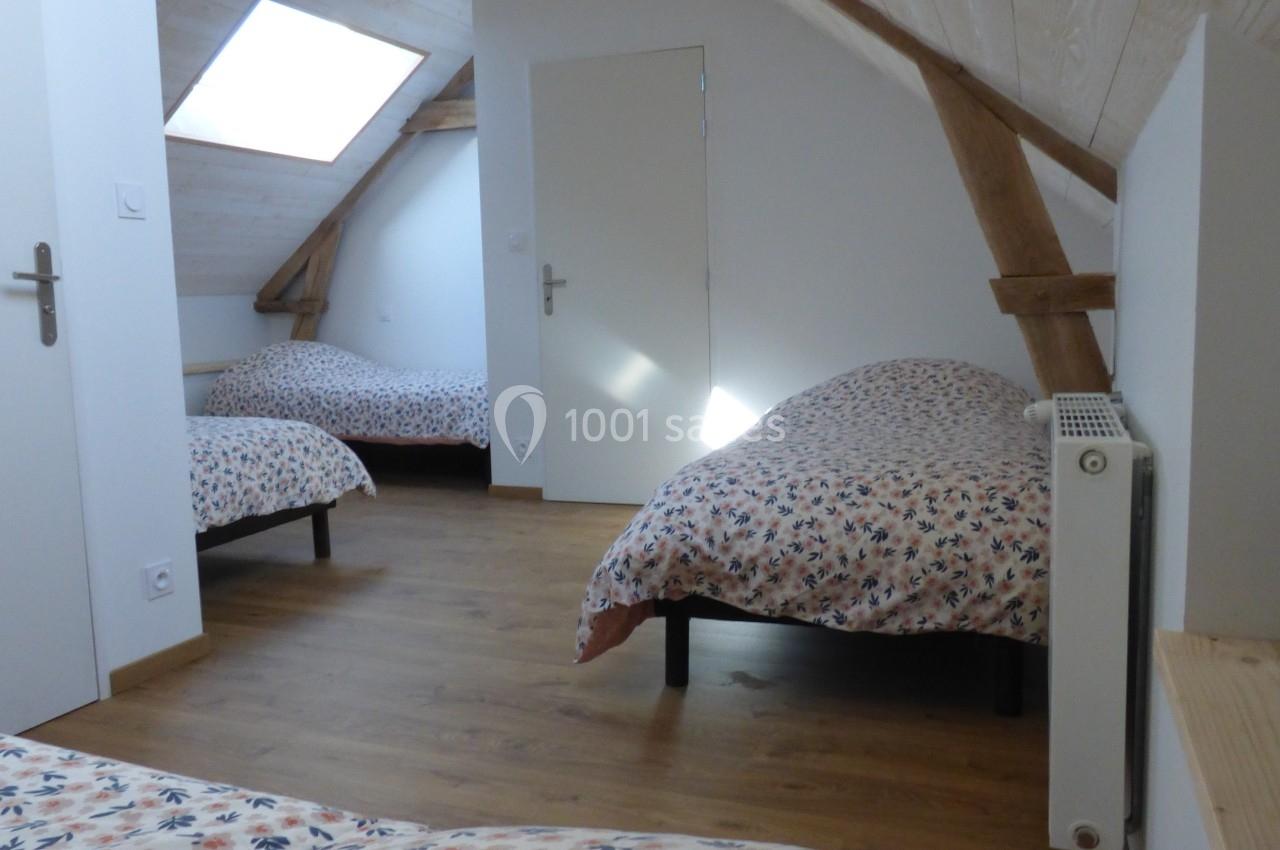 Chambre mansardée avec trois lits simples, parquet au sol, poutres apparentes et lumière naturelle provenant d'une fenêtre…