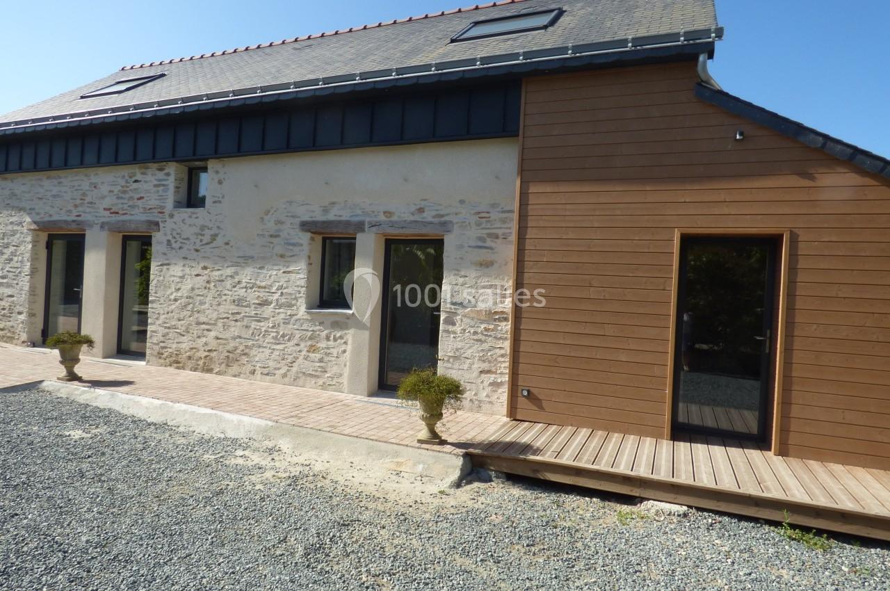 Façade d'une maison rénovée avec murs en pierre, bardage en bois et grandes baies vitrées, entourée de gravier.