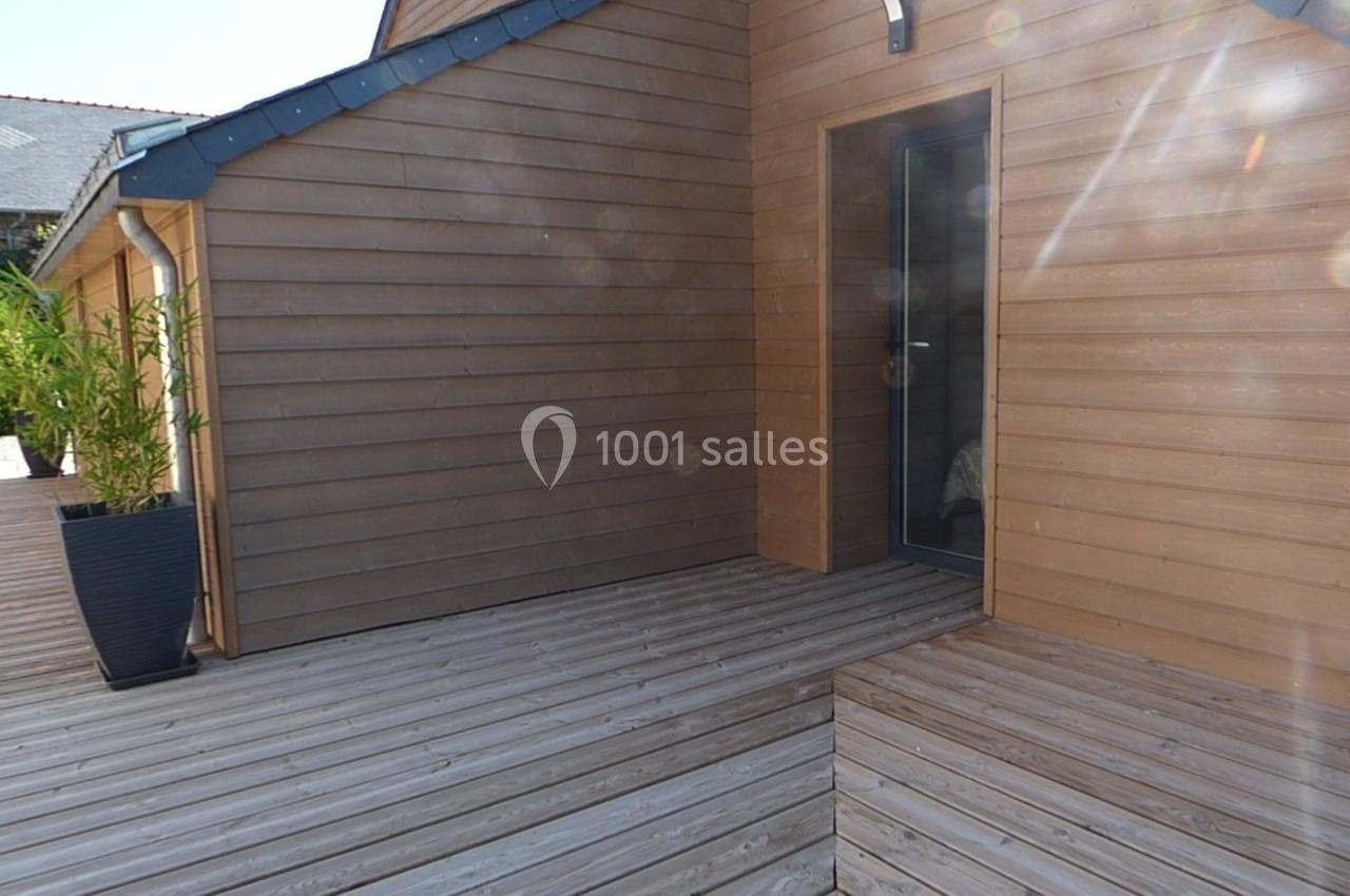 Terrasse en bois attenante à une maison avec bardage en bois, porte vitrée et végétation en arrière-plan.