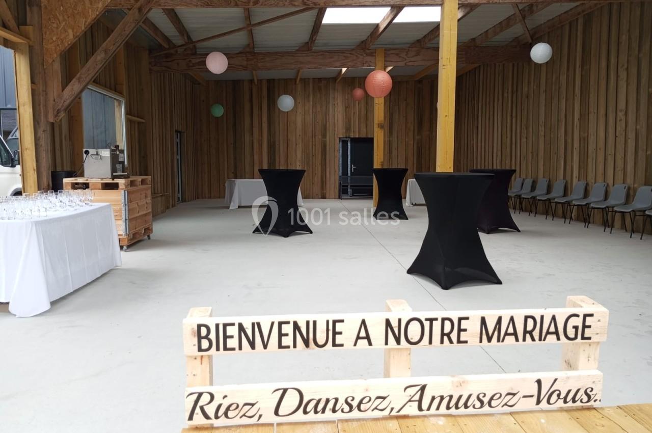 Salle de réception en bois décorée pour un mariage, avec tables hautes, guirlandes et un panneau de bienvenue.