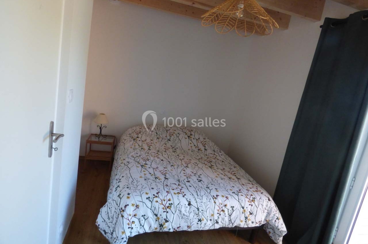 Chambre avec lit double, couverture à motifs végétaux, lampe de chevet, plafond en bois et rideaux sombres.