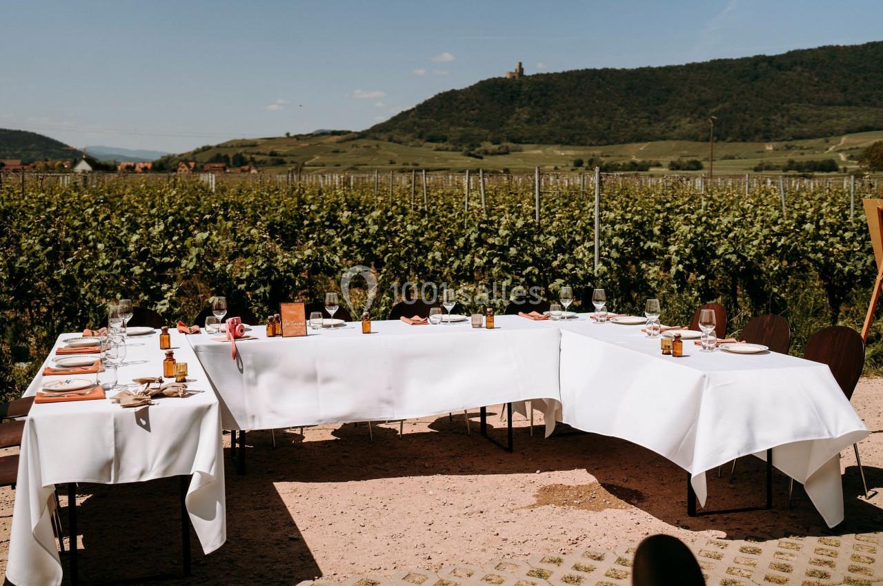 Table dressée en extérieur avec nappes blanches, entourée de vignes et d'une colline en arrière-plan.