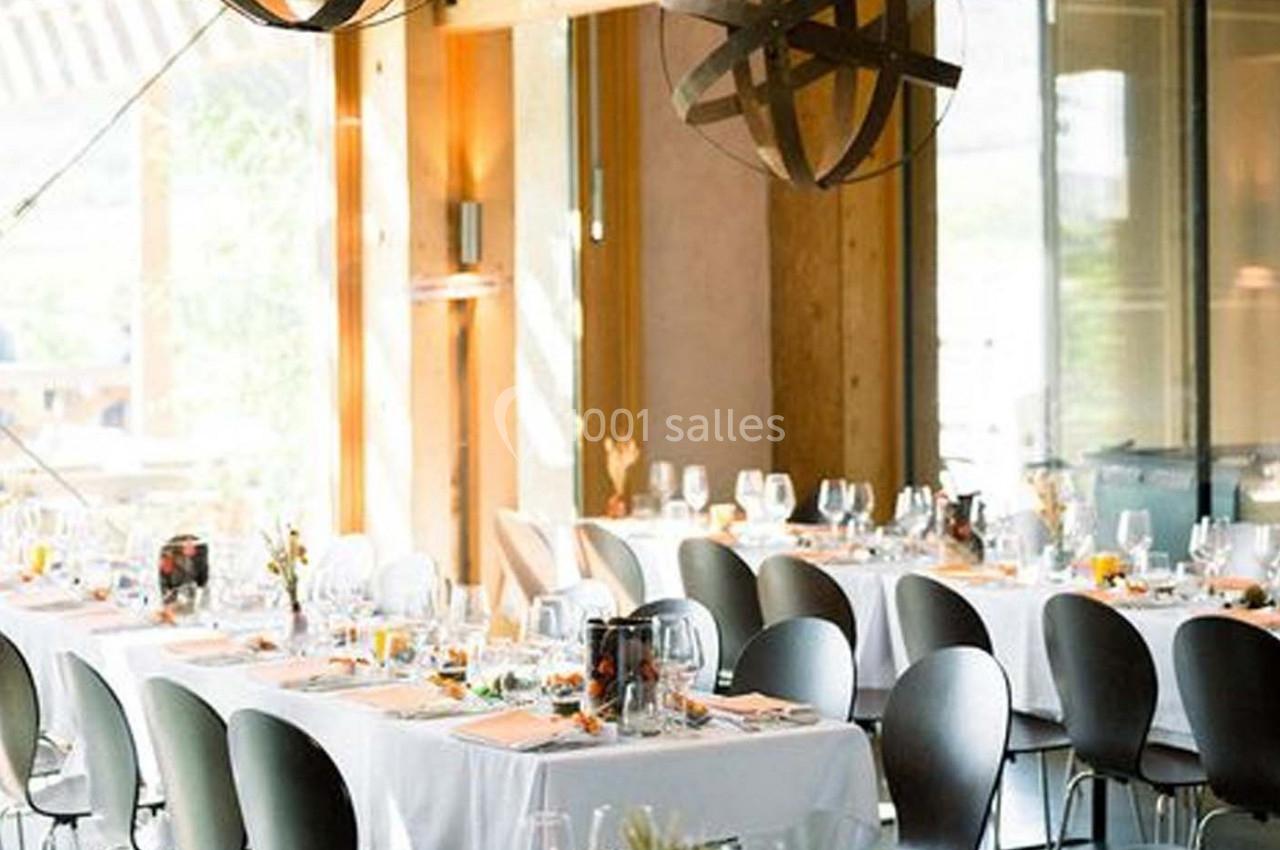 Salle lumineuse avec tables dressées, nappes blanches, chaises noires et décorations élégantes pour un événement.