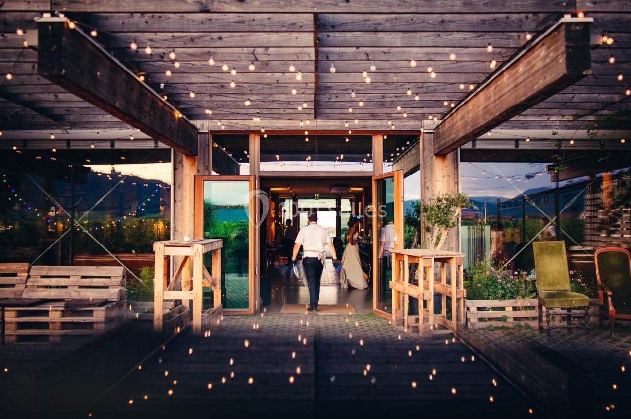 Terrasse en bois décorée de guirlandes lumineuses, avec deux personnes entrant dans un bâtiment vitré.