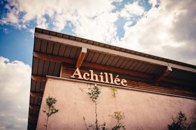 Façade d'un bâtiment en bois et crépi rose avec le mot ’Achillée’, sous un ciel partiellement nuageux.