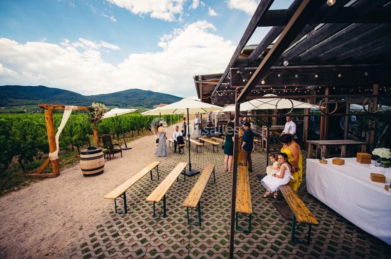 Terrasse extérieure avec bancs en bois, invités rassemblés près d'une arche de mariage, entourée de vignes et collines.