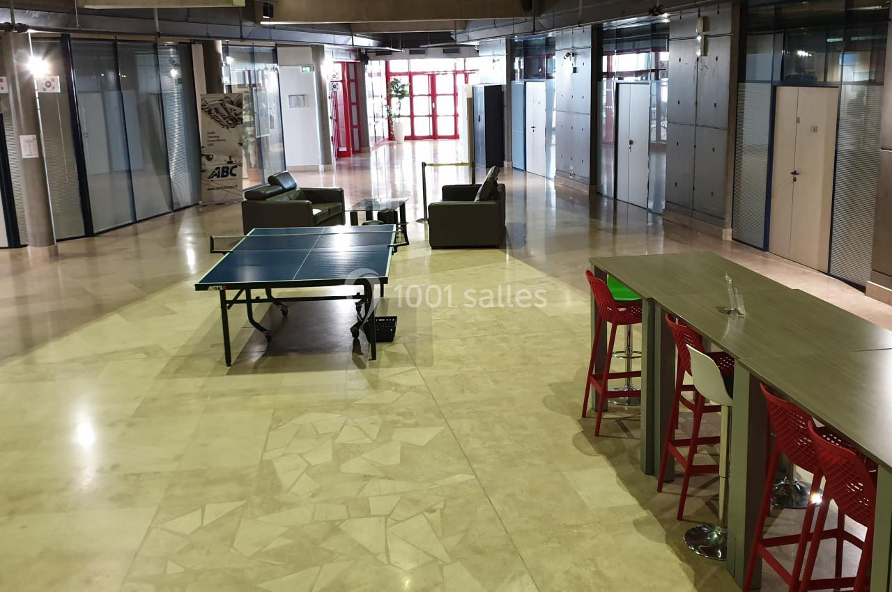 Espace intérieur avec table de ping-pong, comptoir avec tabourets rouges et canapés, entouré de bureaux vitrés.