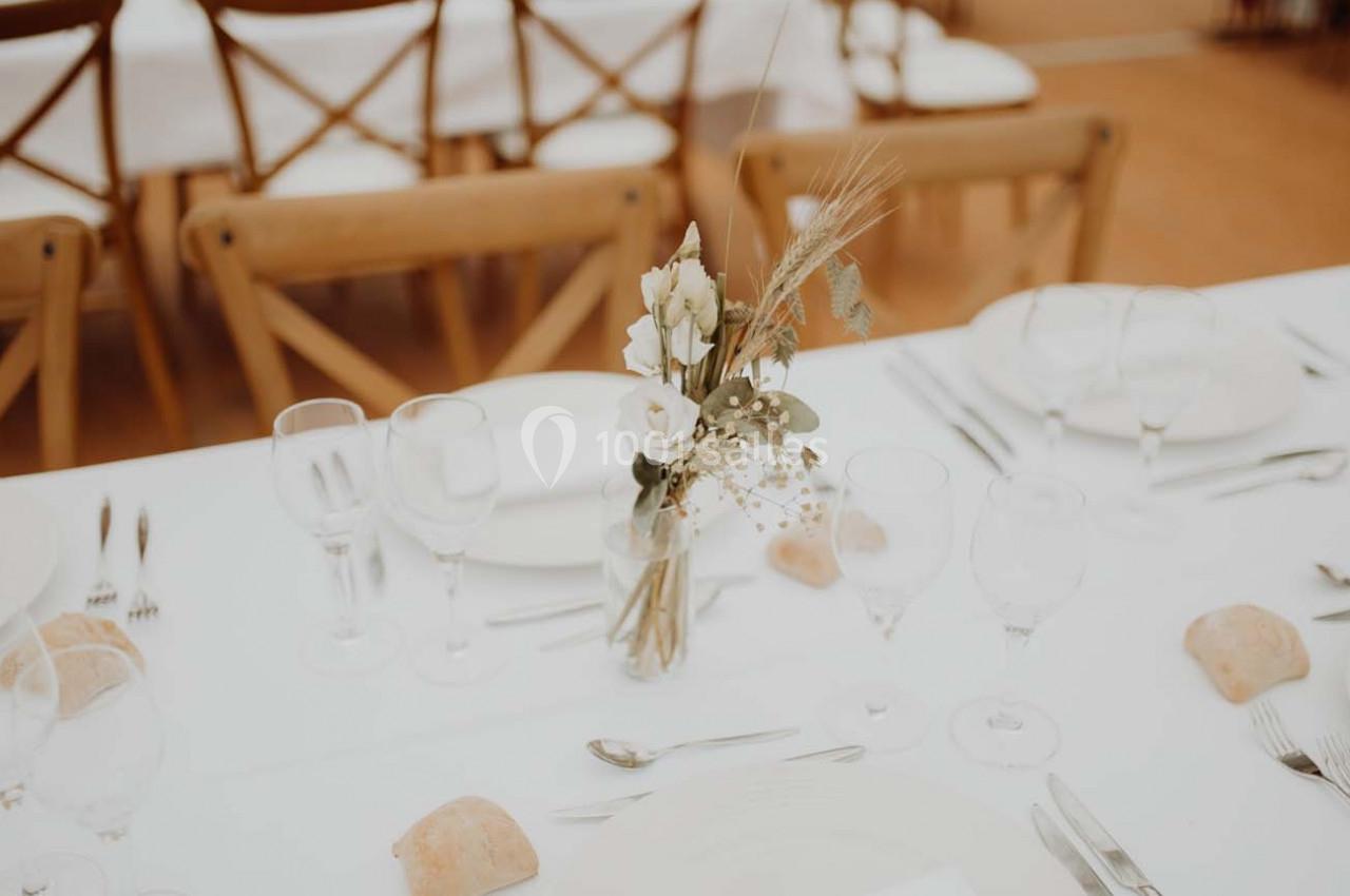 Table dressée avec nappes blanches, vaisselle, verres, couverts et un bouquet de fleurs séchées au centre.