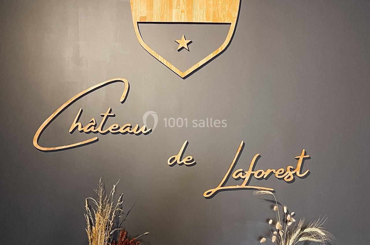 Logo en bois du Château de Laforest sur un mur sombre, entouré de deux vases avec des compositions végétales.