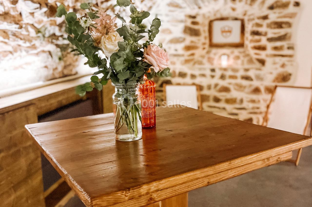 Table haute en bois avec un vase de fleurs et une bouteille rouge, dans une pièce aux murs en pierre apparente.