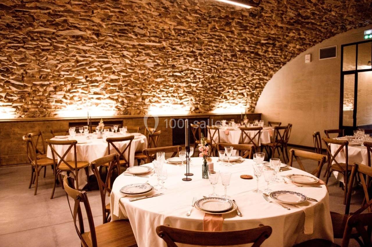 Salle voûtée en pierre avec tables rondes dressées pour un repas, décorée de bougies et vaisselle élégante.