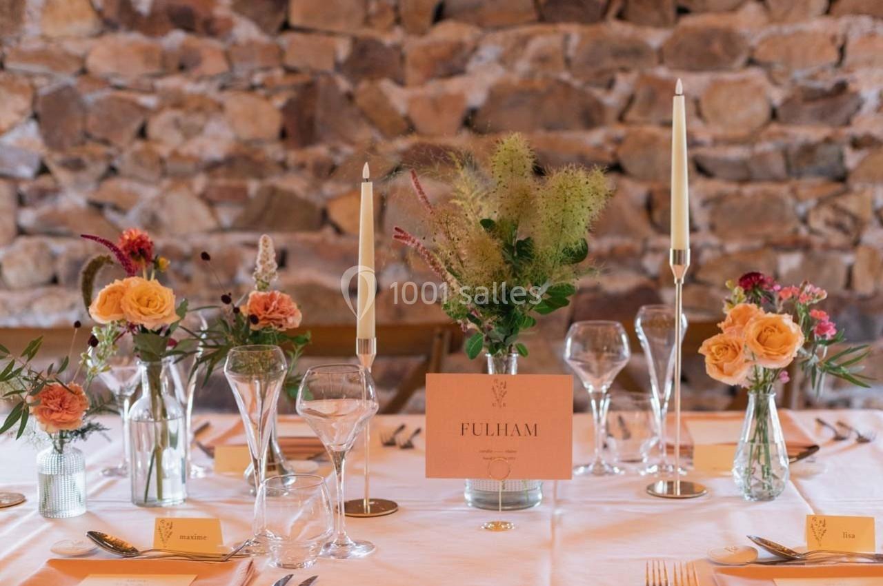 Table élégamment dressée avec fleurs, bougies, verres et un menu ’Fulham’ sur fond de mur en pierres apparentes.