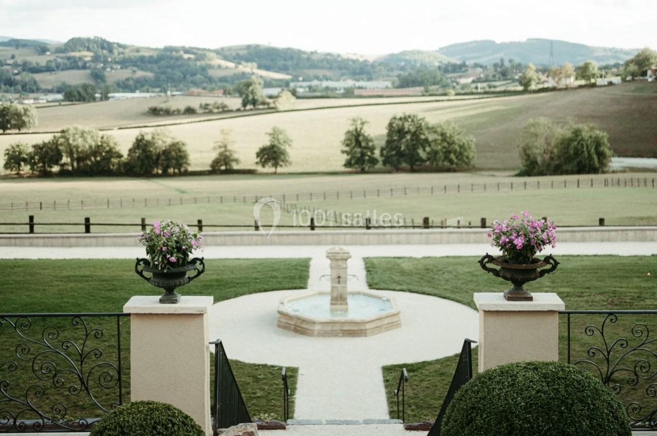 Vue sur un jardin avec une fontaine centrale, encadré par des balustrades et des collines verdoyantes en arrière-plan.