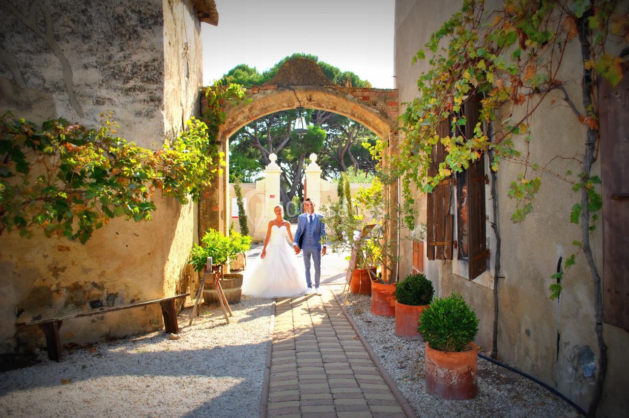 Un couple en tenue de mariage marche dans une allée bordée de plantes et de murs anciens sous une arche en pierre.