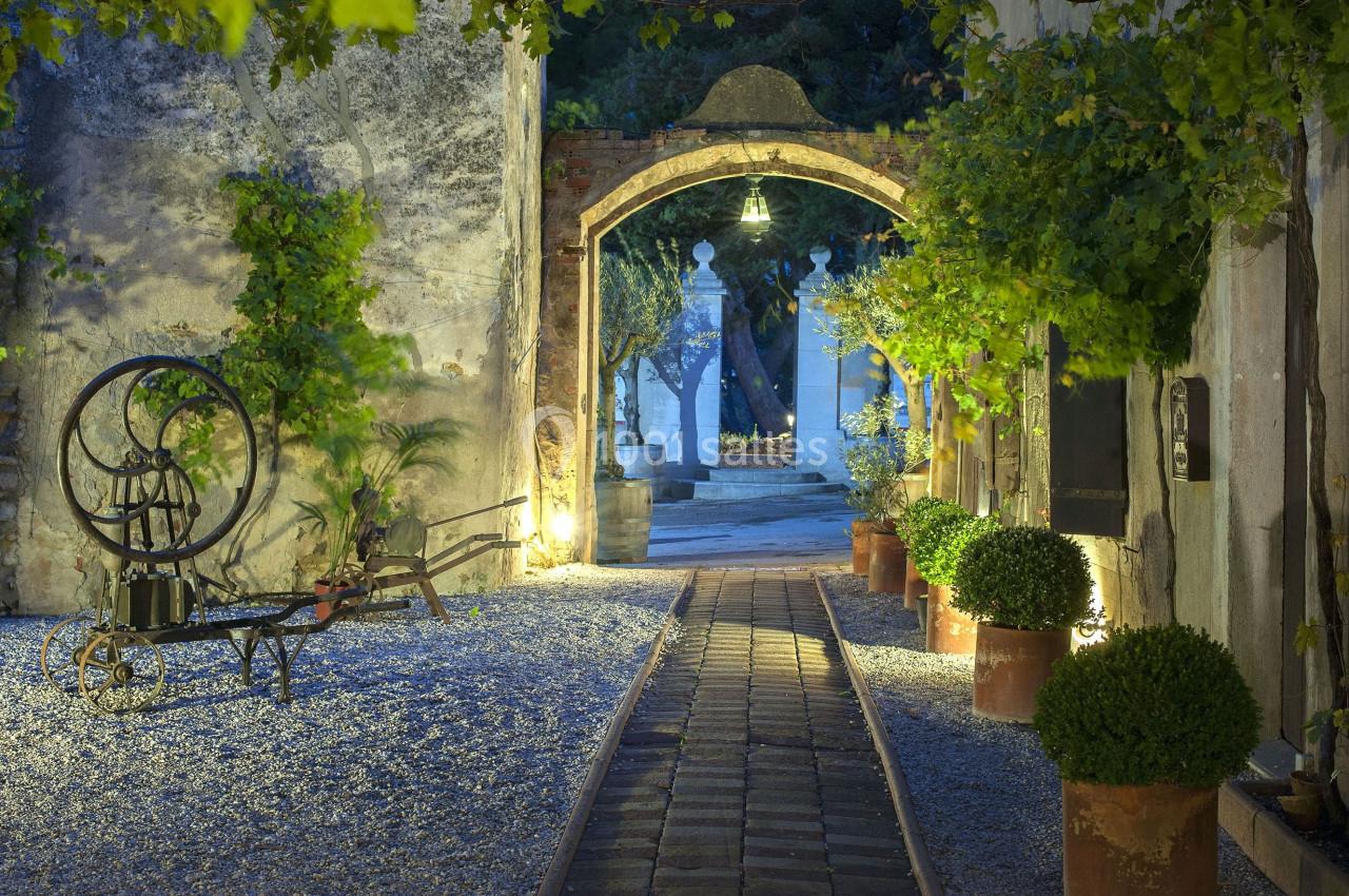 Allée pavée éclairée traversant une arche en pierre, entourée de végétation et de pots décoratifs.