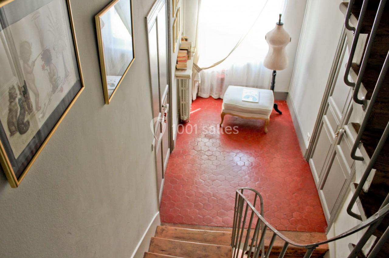 Vue d'un escalier en bois menant à un couloir avec sol en tomettes rouges, un mannequin et une fenêtre lumineuse.
