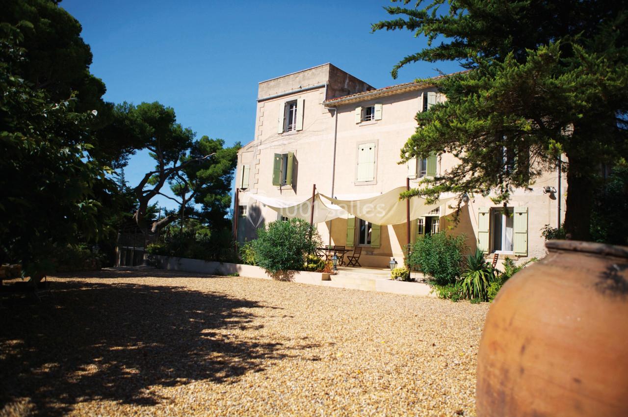 Grande maison en pierre avec volets verts, terrasse ombragée par des toiles, entourée de végétation et d'une cour…